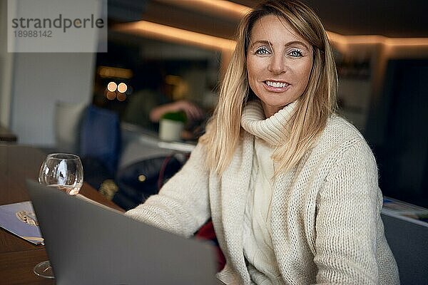 Attraktive blonde Frau  die an einem Laptop arbeitet und in die Kamera lächelt  während sie ein Glas Wein in einer Kneipe oder einem Restaurant genießt