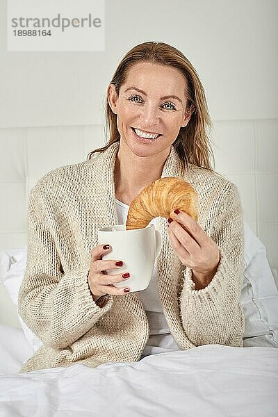 Porträt einer schönen Frau  die beim Frühstück im Bett mit einer Tasse Kaffee oder Milch und einem leckeren französischen Croissant lächelt