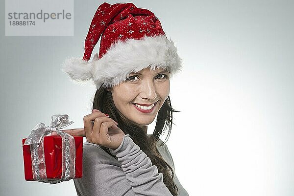 Schöne junge Frau mit Weihnachtsmannmütze und einem dekorativ verpackten Geschenk  das von ihrem Finger über ihre Schulter baumelt  lächelt fröhlich in die Kamera und bietet Platz für Ihren Weihnachtsgruß