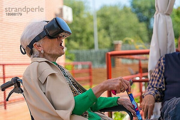 Eine ältere Frau mit der Krankenschwester  die durch eine Virtual Reality Brille im Garten eines Pflegeheims schaut  eine VR Brille mit offenem Mund