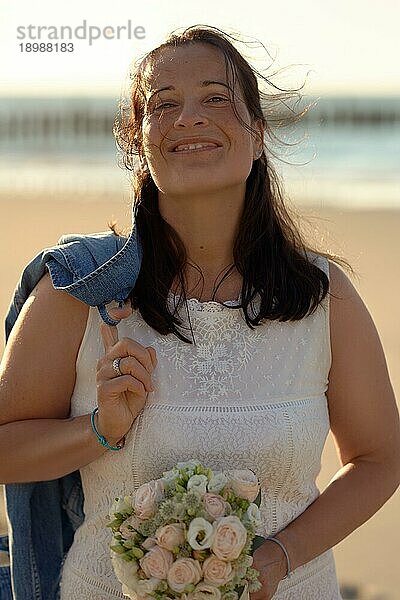 Glückliche moderne Frau in einem eleganten weißen Kleid mit einer Jeansjacke über der Schulter  die lächelnd in die Kamera schaut  während sie einen Brautstrauß an einem tropischen Strand bei Sonnenuntergang trägt  in einem Konzept für Liebe und Ehe