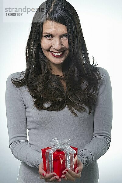 Schöne lächelnde Frau  die einem geliebten Menschen ein Geschenk überreicht und dabei eine romantische rote Geschenkverpackung in den Händen hält  Studioporträt