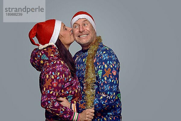 Zärtliches Paar mittleren Alters in festlicher  weihnachtlicher Kleidung  das sich umarmt und küsst  während es Weihnachten zusammen vor einem grauen Hintergrund in Seitenansicht feiert