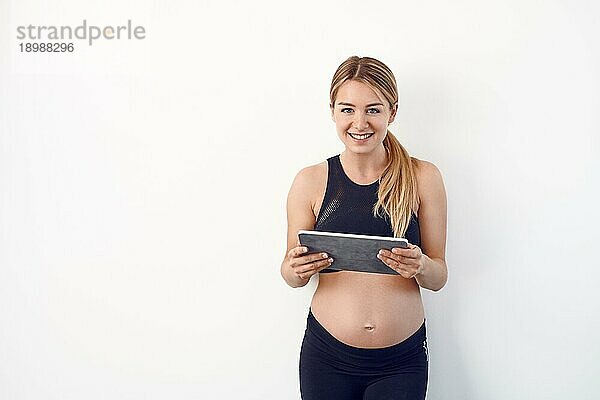 Glückliche junge schwangere Frau mit nacktem Babybauch  die mit einem zufriedenen Lächeln auf einem Tablet PC liest  vor einem weißen Studiohintergrund mit Kopierraum