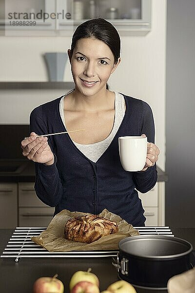 Lächelnde  glückliche Hausfrau beim Backen in der Küche  die über einem frisch gebackenen Apfelkuchen steht  der auf einem Gestell abkühlt  mit einer Tasse Kaffee in der Hand