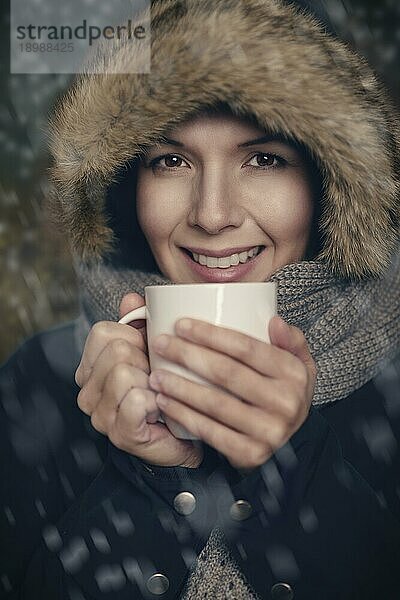 Hübsche junge Frau in warmer Wintermode  warm eingepackt in einen dicken Mantel  einen Strickschal und eine Pelzmütze  die mit einem genussvollen Lächeln einen Becher mit heißer Suppe oder Kaffee in der Hand hält