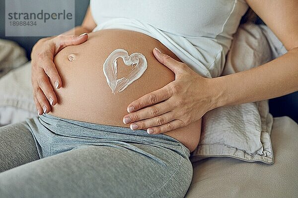 Junge schwangere Frau  die ein Herz in Feuchtigkeitscreme auf ihren Babybauch zeichnet  während sie ihren Bauch in ihren Händen wiegt und sich auf einem Bett in einer Nahaufnahme entspannt