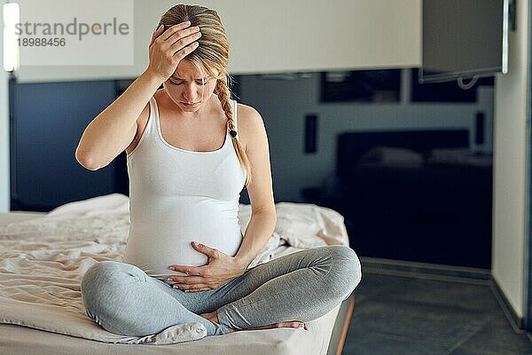 Besorgte hochschwangere junge Frau  die im Schneidersitz auf einem Bett sitzt  ihren geschwollenen Bauch stützt und sich an den Kopf faßt und mit ernster Miene nach unten schaut