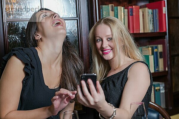 Zwei elegante Frauen  die lachend eine Textnachricht auf ihrem Mobiltelefon lesen  während sie in einer Hotelbibliothek mit hölzernen Bücherregalen zusammenstehen und einen Abend zusammen genießen