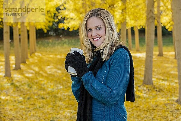 Ein schönes blondes Modell trinkt ein heißes Getränk  während es im Freien in einem Feld mit gelben Blättern posiert