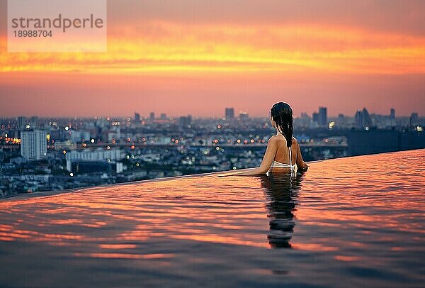 Junge Frau entspannen im Schwimmbad auf dem Dach während erstaunlichen Sonnenuntergang und genießen Stadtbild. Sommerurlaub und Ferien Konzept