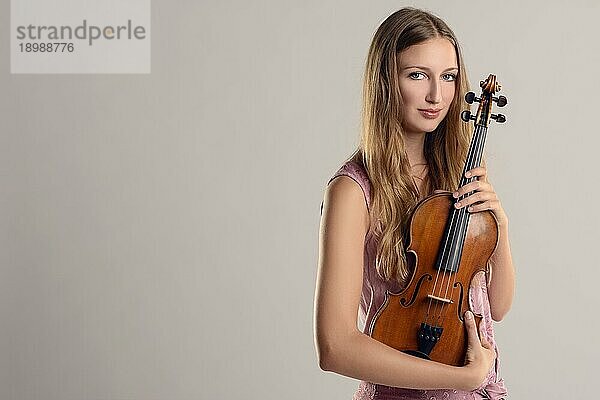 Attraktive junge Musikerin  die ihre Geige im Arm hält und lächelnd in die Kamera schaut  vor einem grauen Hintergrund