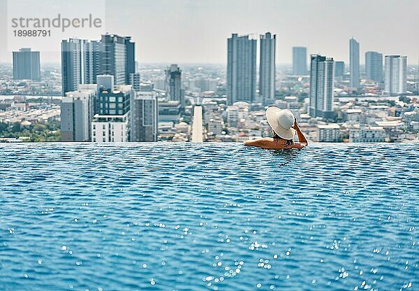 Rückenansicht der jungen Frau in Hut entspannend im Schwimmbad auf dem Dach des Hotels und genießen Stadtbild. Sommerurlaub  Luxuskonzept