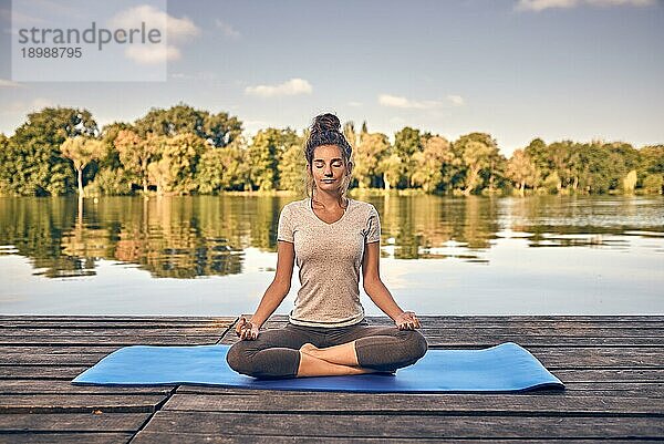 Lächelnde Frau  die auf einer Yogamatte im Lotussitz sitzt und auf einer Holzterrasse oder einem Steg mit Blick auf einen ruhigen See meditiert  im Rahmen eines Konzepts für einen gesunden Lebensstil