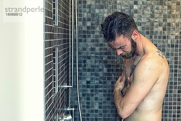 Freundlicher junger Mann mit Vollbart  der sich unter der Dusche einseift und in die Kamera lächelt  Oberkörper mit Kopierraum