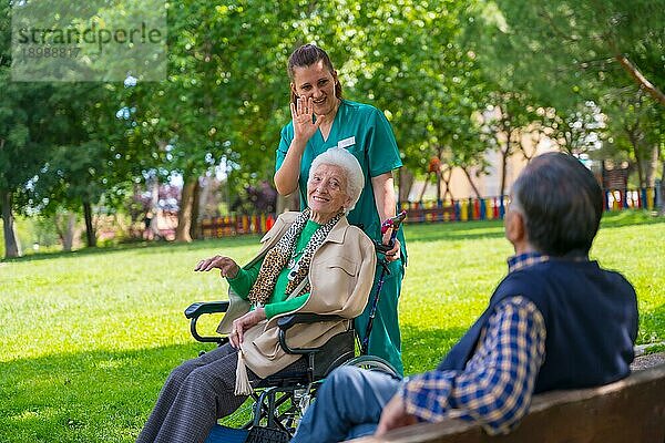 Eine ältere Frau mit der Krankenschwester bei einem Spaziergang durch den Garten eines Pflegeheims im Rollstuhl und beim Begrüßen eines älteren Mannes