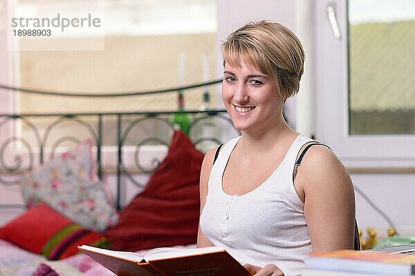 Nahaufnahme Glückliche junge Frau mit kurzen blonden Haaren  die vor ihrem Bett sitzt und ein Buch hält  während sie in die Kamera schaut