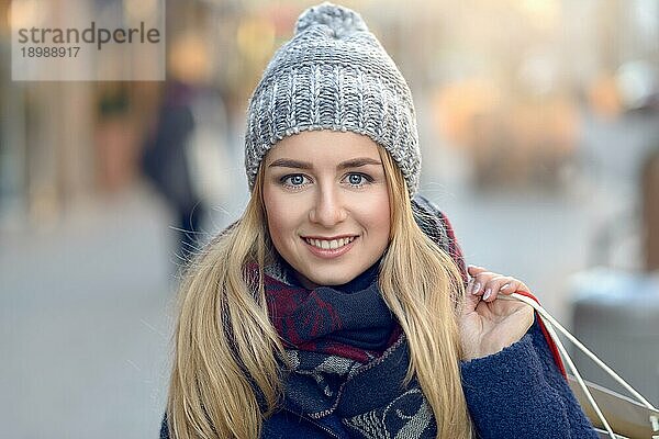 Wunderschöne junge Frau mit Weihnachtseinkäufen in einer gestrickten Wintermütze  die fröhlich lächelt  während sie auf einer städtischen Straße hinter sich blickt