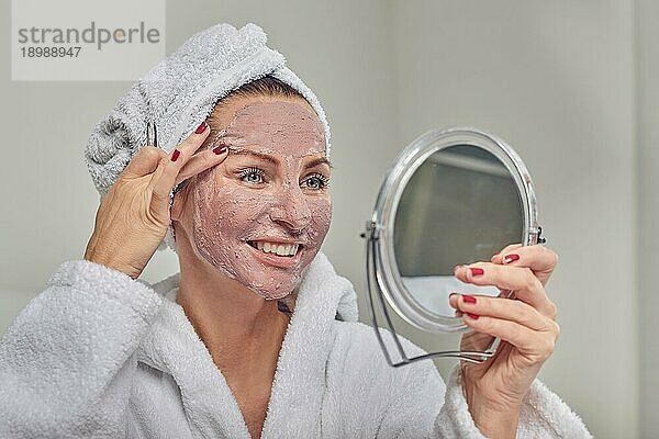 Attraktive Frau mit Gesichtsmaske und Schönheitsbehandlung  die einen Spiegel hält  während sie ihre Augenbrauen mit einer Pinzette zupft