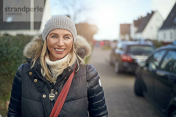 Lächelnde  glückliche Frau mittleren Alters  die an einem kalten Wintertag in einer warmen Jacke und Strickmütze in einer Vorstadtstraße im Freien steht