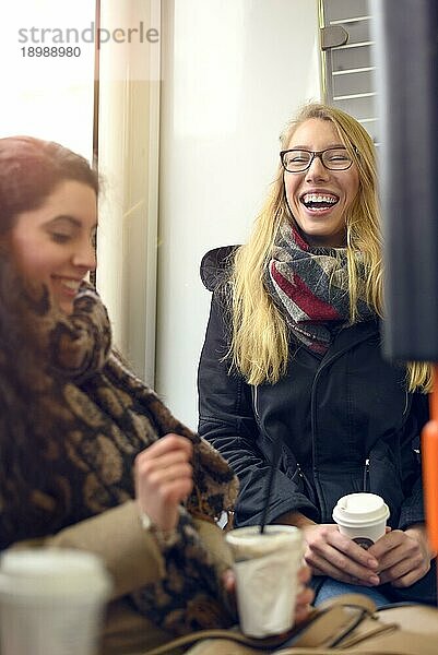 Zwei sitzende blonde und brünette Freundinnen in Mänteln und mit Kaffee in der Hand teilen einen humorvollen Moment