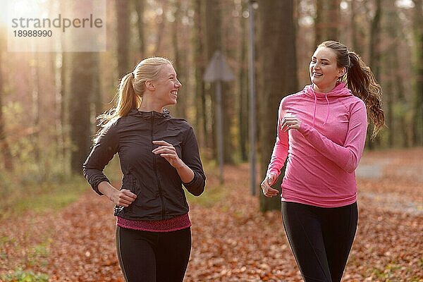 Zwei attraktive  fitte junge Damen joggen gemeinsam durch einen herbstlichen Wald und unterhalten sich dabei  während sie durch die Bäume laufen  ein Konzept für gesunden Lebensstil und Sport