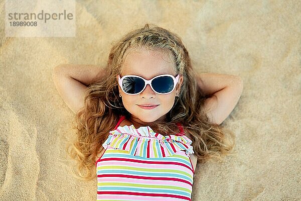 Draufsicht auf hübsches kleines blondes Mädchen mit Sonnenbrille am Sandstrand liegend während der Sommerferien. Meer entspannen  glückliche Kindheit Konzept