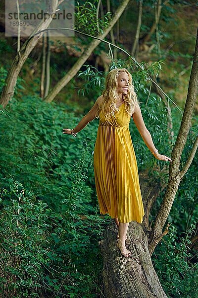Unbeschwerte barfuß attraktive blonde Frau balanciert auf einem alten umgestürzten Baumstamm in einem Wald und trägt ein elegantes gelbes Kleid  das mit einem glücklichen freundlichen Lächeln zur Seite schaut