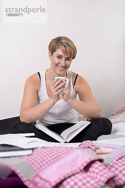 Hübsche junge Studentin mit einem charmanten  fröhlichen Lächeln  die im Schneidersitz mit ihren Lehrbüchern und einer Tasse Tee oder Kaffee auf ihrem Bett sitzt und eine Pause vom Lernen macht