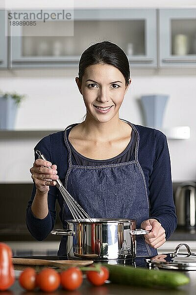 Fröhliche  freundliche junge Hausfrau  die am Herd steht und einen Schneebesen über einen Topf hält  während sie das frische Gemüse für das Abendessen zubereitet und der Kamera ein schönes Lächeln schenkt