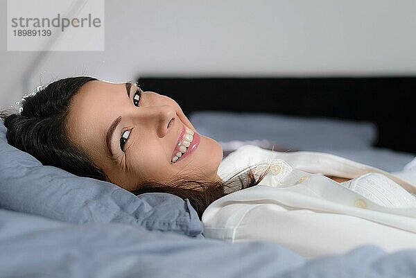 Nahaufnahme einer erfrischten  glücklichen jungen Frau  die im Bett liegt und den Kopf in die Kamera dreht