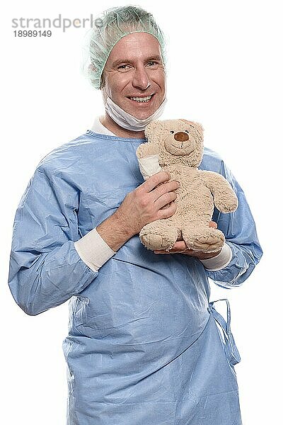 Freundlich lächelnder Kinderarzt mittleren Alters in blauem Kittel  der einen Teddybär mit bandagiertem Arm hält  um einen jungen Patienten zu beruhigen  vor weißem Hintergrund