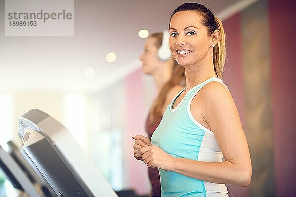 Attraktive Frau mittleren Alters in einem mintfarbenen Top steht auf dem Laufband im Fitnessstudio  schaut in die Kamera und lächelt mit Kopie Raum