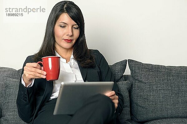 Attraktive  stilvolle  berufstätige Frau  die sich mit einer Tasse Kaffee in der Hand auf einem Sofa ausruht und auf einem Tablet PC für Freizeit  Studium oder geschäftliche Kommunikation über das Internet liest