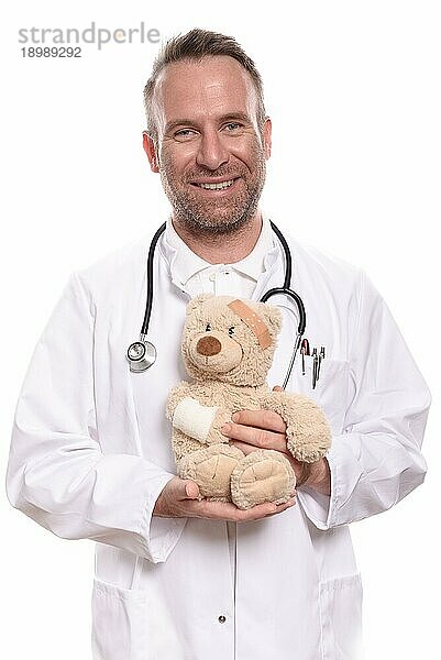 Freundlich lächelnder Kinderarzt mittleren Alters mit unrasierten Bartstoppeln  der einen Teddybär mit bandagiertem Arm hält  um einen jungen Patienten zu beruhigen  vor weißem Hintergrund