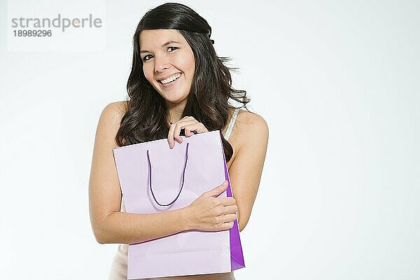 Begeisterte junge Frau  die ihre Einkäufe in einer hübschen lila Einkaufstasche besitzergreifend an ihre Brust drückt  während sie glücklich lächelt und ihre Zufriedenheit zeigt  vor weißem Hintergrund