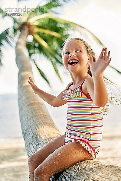 Glückliche kleine blonde Mädchen spielen am Strand sitzen auf Palme. Sommerurlaub  Kindheit und Spaß Konzept
