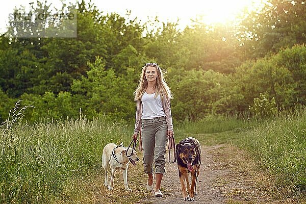 Glückliche junge Frau  die mit ihren Hunden auf einem grasbewachsenen Feldweg im Frühling spazieren geht und in die Kamera schaut