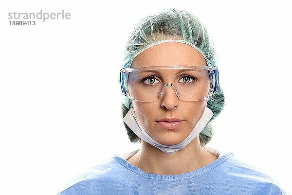 Junge Krankenschwester oder Ärztin in OP Kleidung mit OP Kittel  Maske  Schutzbrille und Kappe  die direkt in die Kamera blickt  Kopf und Schultern vor weißem Hintergrund