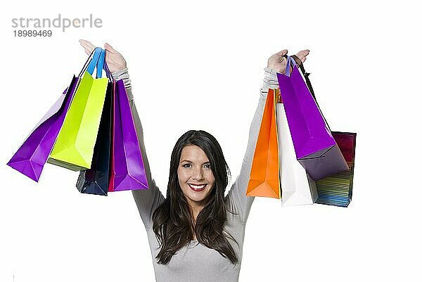 Fröhliche Einkäuferin  die mit einem strahlenden Lächeln eine Sammlung bunter Einkaufstaschen hochhält und sich über ihre erfolgreichen Einkäufe freut  vor weißem Hintergrund