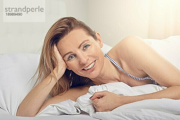 Glückliche  freundliche  hübsche junge Frau  die sich im Bett auf der Seite liegend auf einen Ellbogen stützt und glücklich in die Kamera grinst