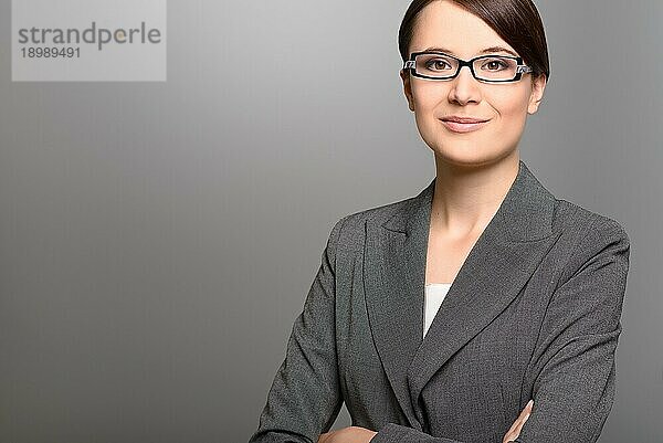 Stilvolle junge Geschäftsfrau mit freundlichem Gesichtsausdruck und Brille  die direkt in die Kamera blickt  Nahaufnahme ihres Gesichts auf grauem Hintergrund mit Leerzeichen