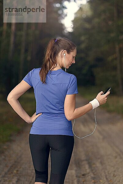 Attraktive glückliche Frau  die mit ihrem MP3 Player und Ohrstöpseln auf einem Feldweg im Wald steht und Musik hört