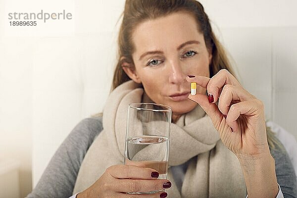 Porträt einer kranken Frau im Bett  die verärgert in die Kamera schaut  während sie ein Glas Wasser und eine Tablette gegen Grippesymptome hält