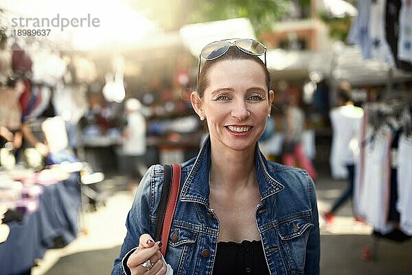 Glückliche  attraktive  stilvolle Frau  die auf einem Markt im Freien einkauft und mit einem breiten  warmen  freundlichen Lächeln in die Kamera schaut