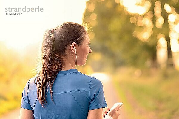 Attraktive glückliche Frau  die mit ihrem MP3 Player und Ohrstöpseln auf einer Landstraße im Sonnenlicht steht und Musik hört