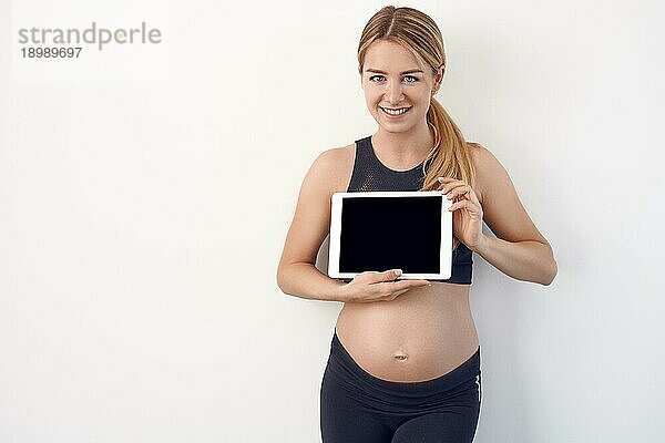 Lächelnde freundliche junge schwangere Frau  die dem Betrachter ein weißes modernes Tablet mit Bildschirm vor einem Studiohintergrund mit Kopierraum entgegenhält