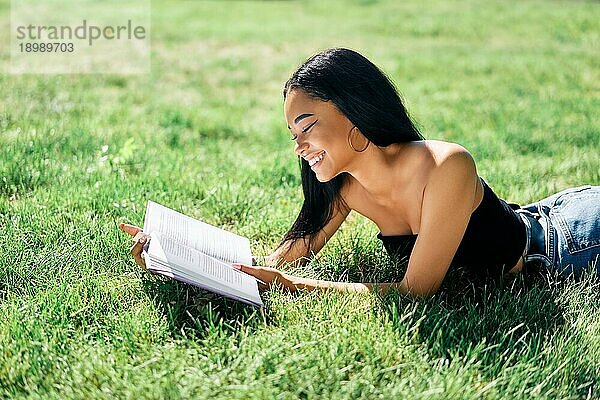 Hübsche lächelnde afroamerikanische Frau  die im Gras liegend im Park ein Buch liest. Entspannen und Hobby Konzept