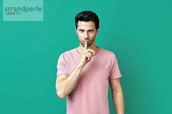 Junger Mann in rosa TShirt hält Finger auf den Lippen  so daß Hush Geste vorblauem Hintergrund. Pst  Stille Konzept
