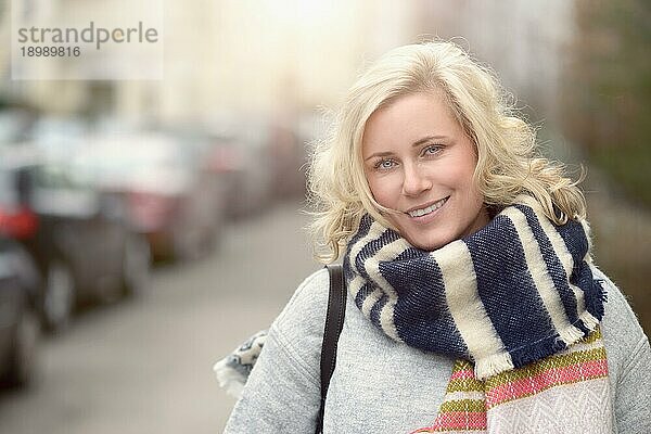 Lächelnde  attraktive  junge  blonde Frau mit Wollschal  die am Rande einer städtischen Straße steht und in die Kamera schaut  Oberkörperansicht